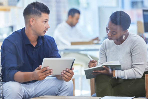 Two colleagues converse seated in an office with notebooks and a tablet