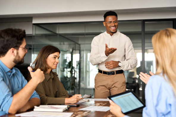 A group of colleagues converse at a conference table and one person is standing to present