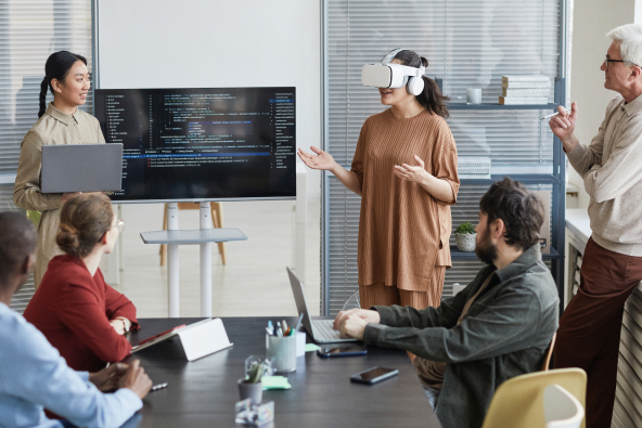 A group of software developers gathered in a meeting room in front of code on a monitor with one person wearing virtual reality goggles