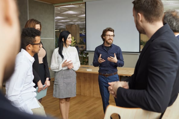 A group of professionally dressed colleagues stand in a circle listening as someone speaks