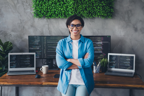 A tech professional leans against a desk with multiple monitors with arms crossed and smiling